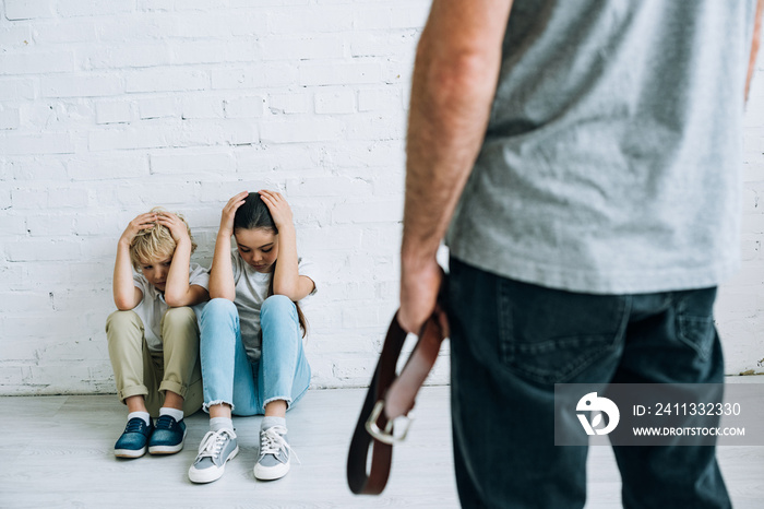 cropped view of abusive father holding belt and scared kids sitting on floor