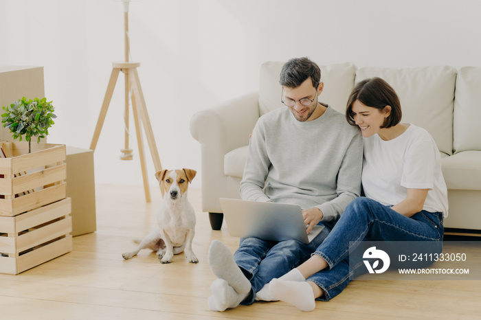 Indoor shot of lovely couple move in new dwelling, use modern laptop for searching design ideas for their flat, sits on floor near sofa, pedigree dog poses near cardboard boxes. Moving Day concept