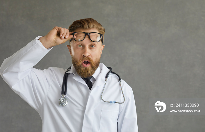 Amazed shocked doctor wearing white coat and stethoscope taking off eyeglasses looking at camera with disbelief emotion on face. Studio shot portrait of staring therapist over grey background