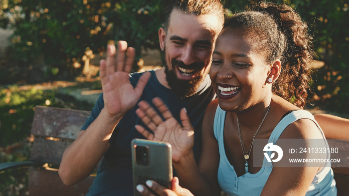 Happy smiling interracial couple taking selfie on phone while sitting on park bench.