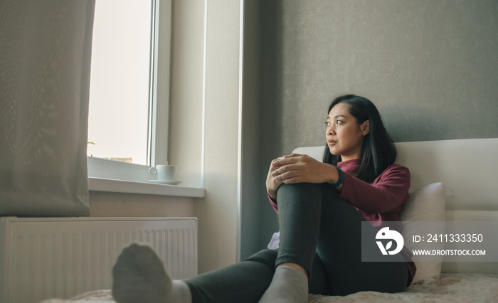 Woman is sitting on the bed looking out to the windows and thinking or missing of someone.