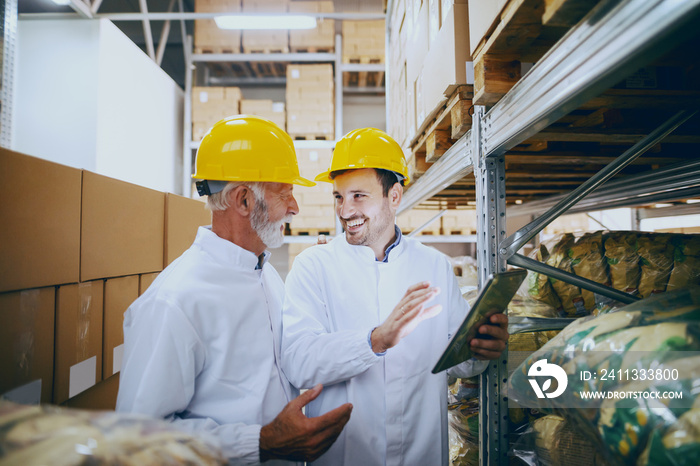 Two smiling Caucasian employees in white uniforms and yellow helmets on heads checking on goods in warehouse. Younger one showing what is on stock by using tablet.