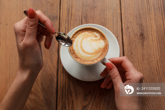 Lonely woman with nice manicure in warm wood sweater drinking coffee in the morning, top view of female hands holding cup of hot beverage on wooden desk, retro toned.