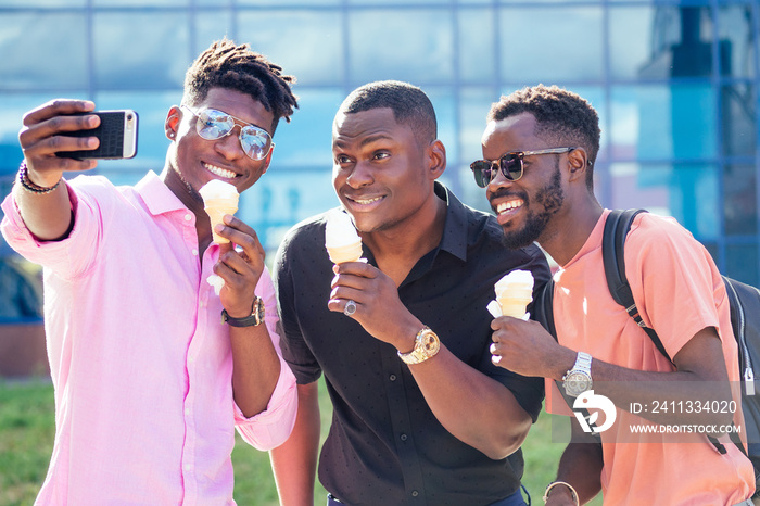 a group of three friends African American guy eating ice cream in a waffle horn and photographing selfie on phone in the summer in the park