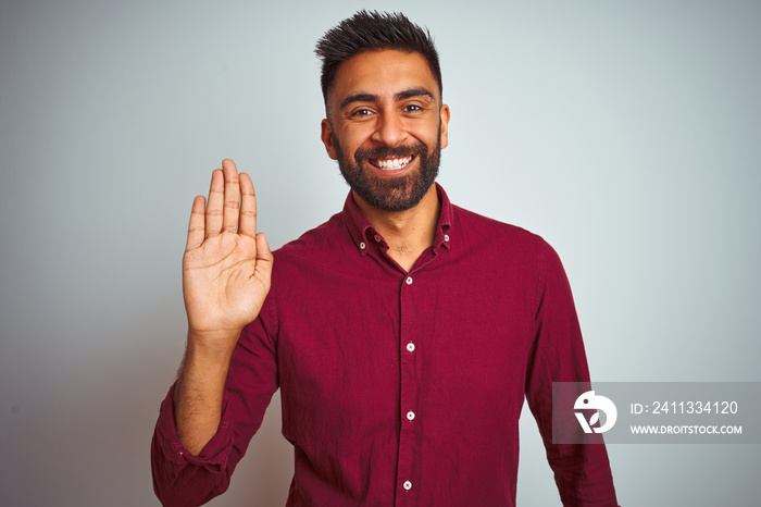 Young indian man wearing red elegant shirt standing over isolated grey background Waiving saying hello happy and smiling, friendly welcome gesture