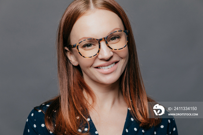 Cheerful close up shot of pretty woman with brown hair, toothy smile, wears spectacles, dressed in formal blouse, poses over grey background, being pleased to meet with colleagues on meeting