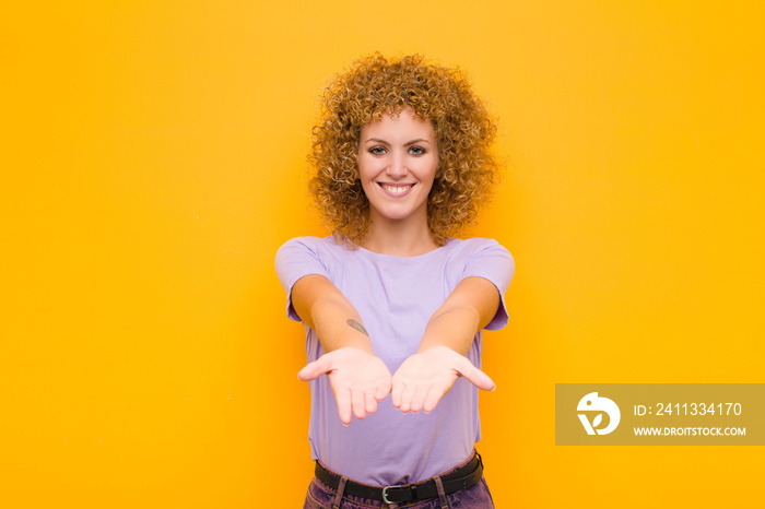 young afro woman smiling happily with friendly, confident, positive look, offering and showing an object or concept against orange wall