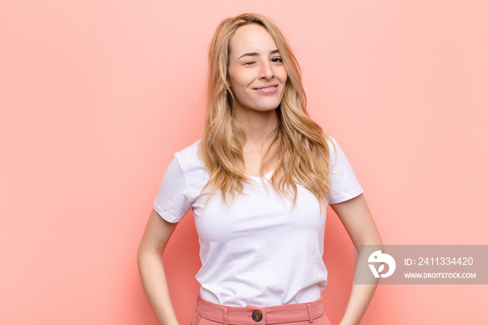 young pretty blonde woman looking happy and friendly, smiling and winking an eye at you with a positive attitude against flat color wall