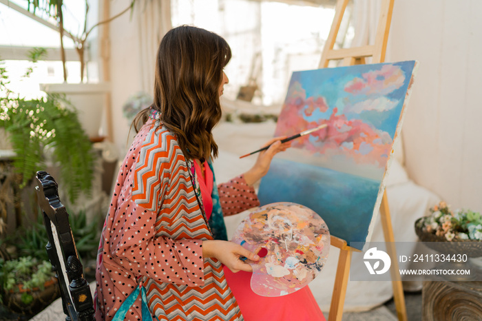 Female artist painting on canvas in her art studio. Wearing elegant boho outfit.