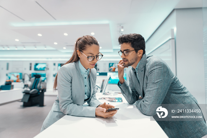 Cute multicultural couple in formal wear discussing about smart phone they want to buy. Tech store interior.