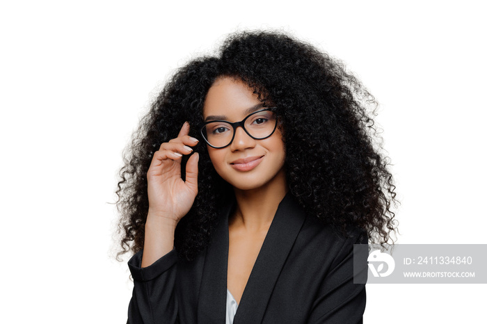 Close up portrait of calm satisfied Afro American woman with bushy curly hairstyle, keeps hand raised, has healthy skin, wears spectacles, black elegant outfit isolated on brown wall. Face expressions