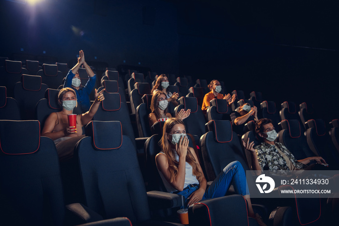 Cinema, movie theatre during quarantine. Coronavirus pandemic safety rules, social distance during movie watching. Men and women wearing protective face mask sitting in a rows of auditorium, eating