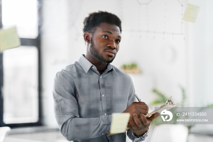 business and people concept - african american businessman with clipboard looking at charts on office glass board