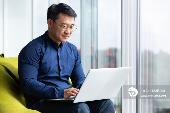 Successful and smiling asian man working with laptop inside office with notebook, senior businessman in glasses and shirt.