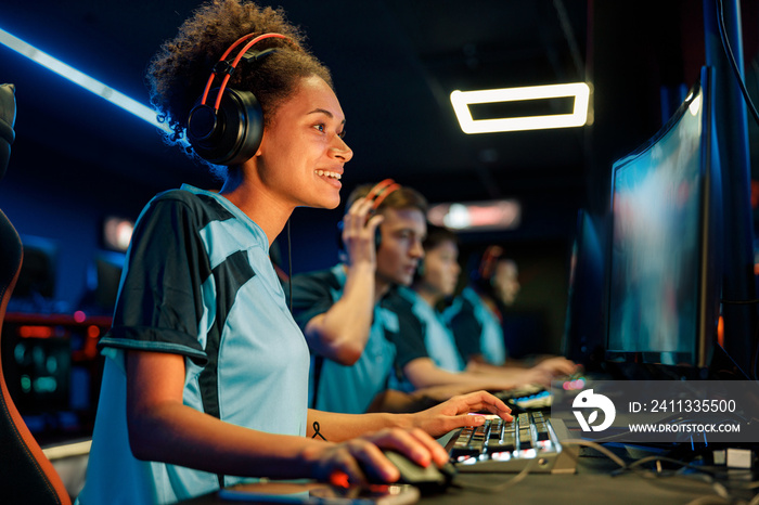 Happy African female cybersport gamer wearing headphones playing online video game while participating in esports tournament in internet cafe