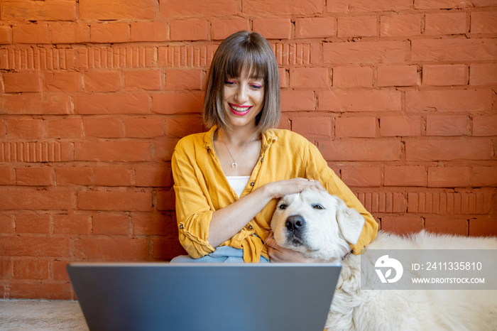 Young woman working on laptop computer while sitting with her cute dog on brick wall background at home. Work from home and friendship with pets concept