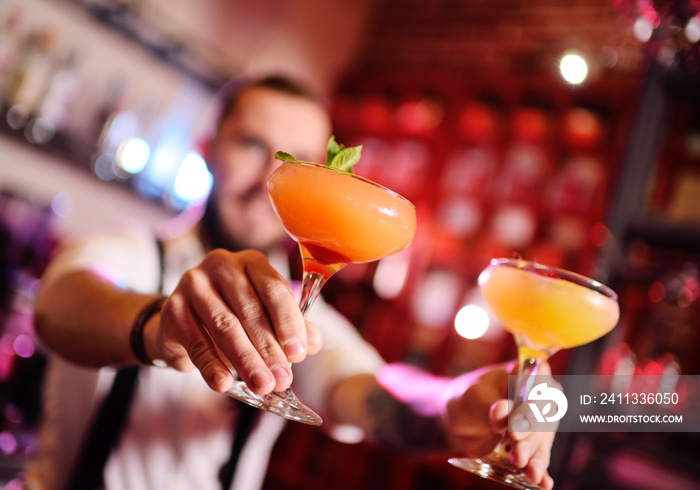 handsome bartender prepares an orange alcoholic cocktail and smiles on the background of a bar or a night club