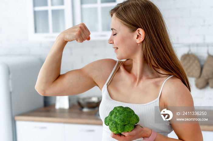 Strong woman with broccoli in the kitchen. Young healthy girl shows her strong hands.