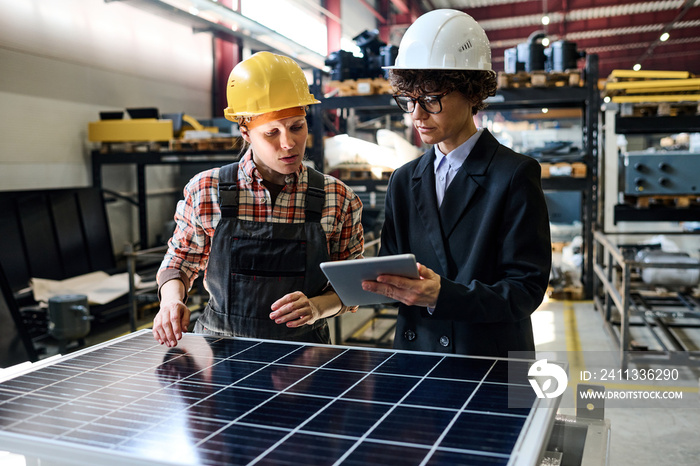 Confident manager with tablet showing young female engineer online data about characteristics of solar panels produced at factory