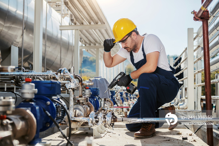 Handsome caucasian worker in overall and helmet on head crouching and using drill while crouching. Oil industry.