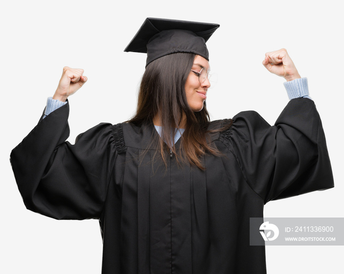 Young hispanic woman wearing graduated cap and uniform showing arms muscles smiling proud. Fitness concept.