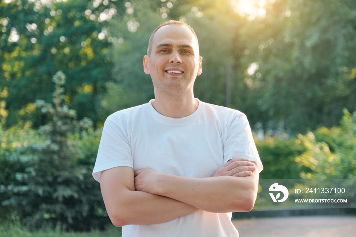 Outdoor portrait of confident smiling middle-aged sports man. Positive handsome male with arms crossed in white t-shirt looking at the camera