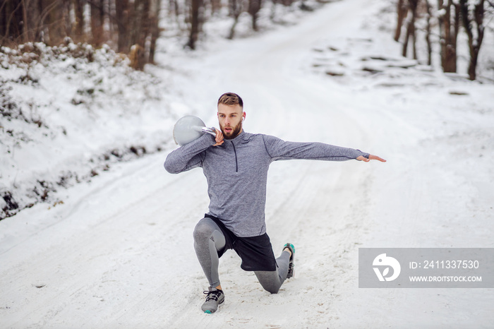 Fit sportsman lifting kettlebell while doing lunges on snowy path in woods. Winter fitness, healthy life, bodybuilding