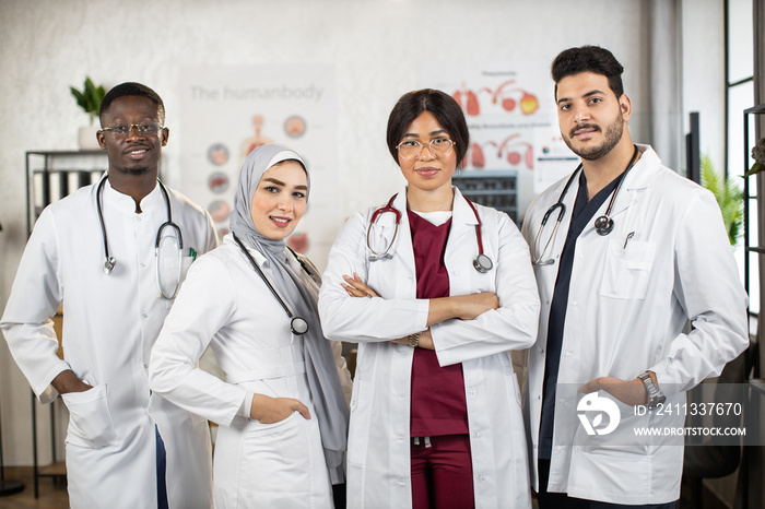 Team of female and male multiracial doctors in white lab coats keeping hands crossed or in the pockets while standing together at medical center. Concept of people, medicine and teamwork.