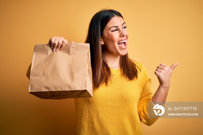 Young beautiful woman holding take away paper bag from delivery over yellow background pointing and showing with thumb up to the side with happy face smiling