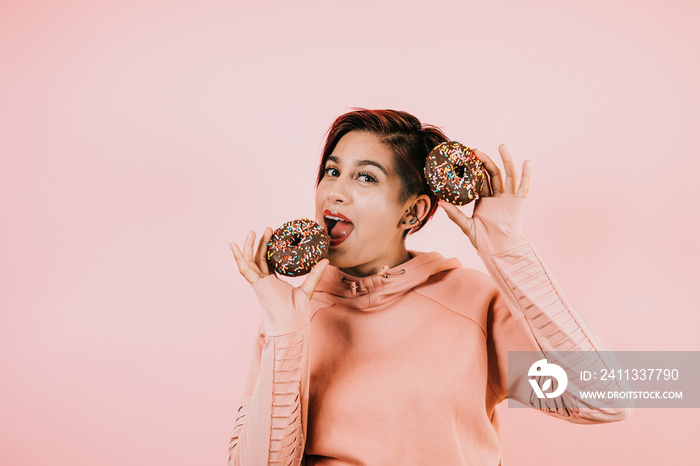 Portrait of young hispanic woman eating chocolate donuts on coral pink background in Mexico Latin America