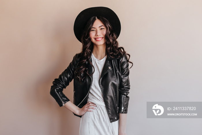 Asian girl in leather jacket standing with hand on hip. Studio shot of smiling chinese woman in hat isolated on beige background.