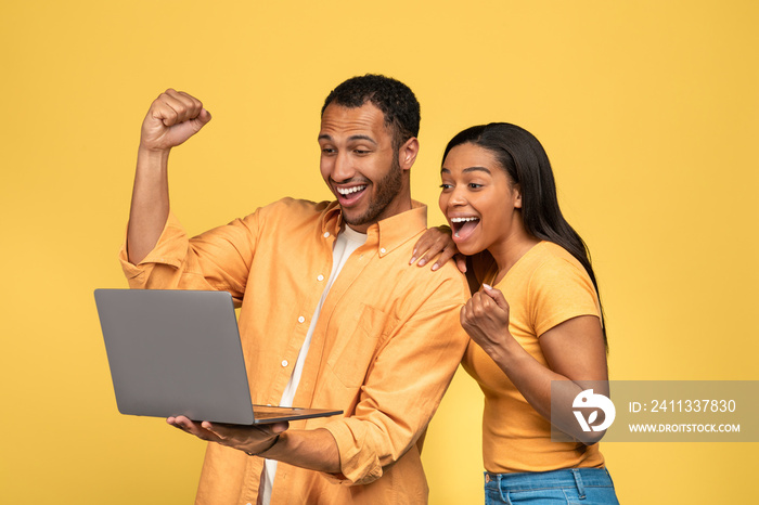 Millennial black couple making YES gesture, looking at laptop, celebrating success or achievement on yellow background