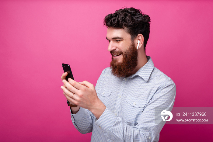 Young bearded man using phone and his earpods on pink background. Wireless technology.