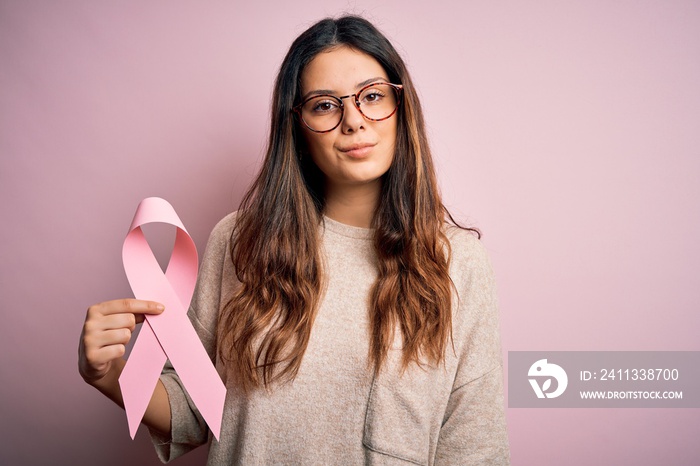 Young beautiful brunette woman wearing glasses holding pink cancer ribbon symbol with a confident expression on smart face thinking serious