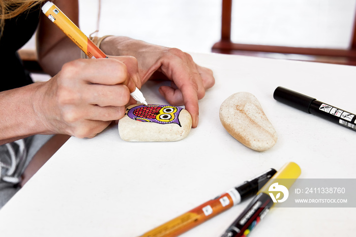 Hands of a woman painting an owl figure on a stone with an acrylic pen. Stone painting handicraft.