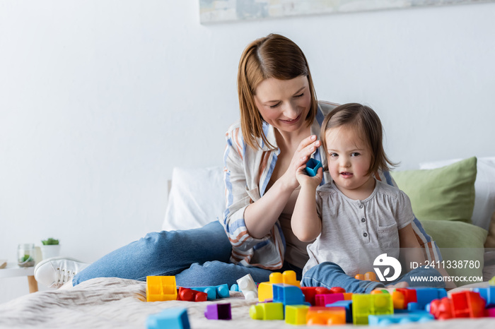 Smiling parent holding building block near daughter with down syndrome on bed.