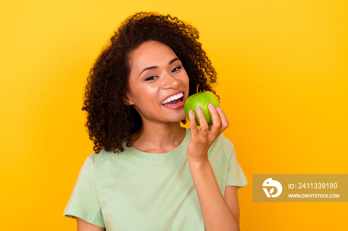 Photo of positive cheerful girl dressed green t-shirt biting white teeth green apple isolated yellow color background