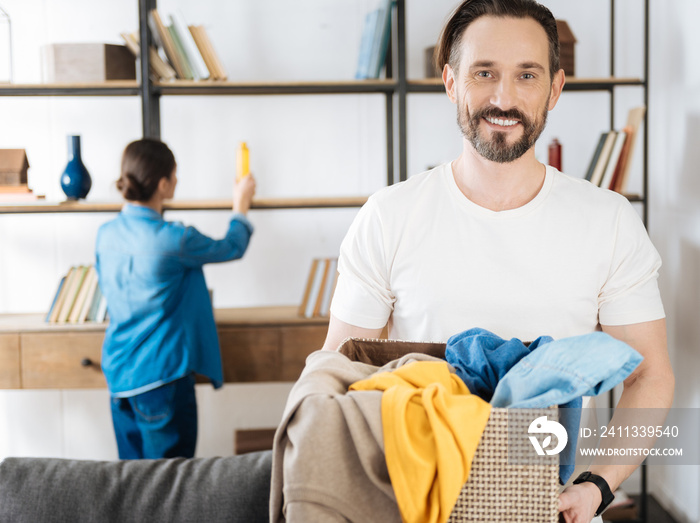 Weekly laundry. Cute pretty optimistic husband standing on the blurred background while looking at the camera and taking basket with clothes