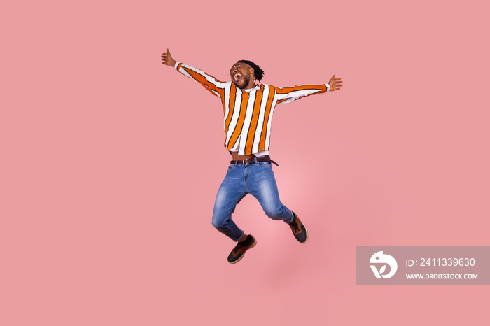 Extremely happy excited afro-american man with dreadlocks in striped shirt highly jumping widely opening hands and screaming feeling freedom. Indoor studio shot isolated on pink background.