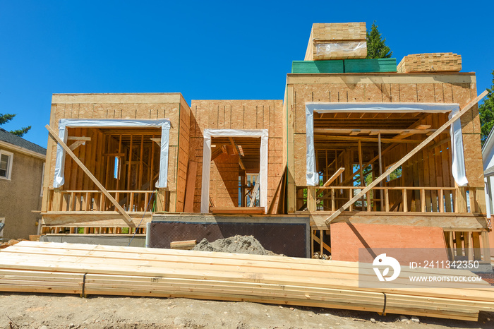 A single family home under construction. The house has been framed and covered in plywood. Stacks of board timber in front and stack of 2x4 boards on the top.