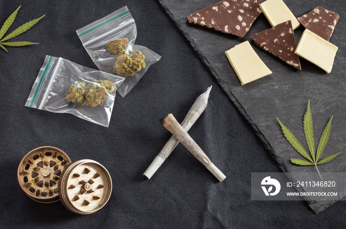 Weed and sweets: joints, marijuana leaves, buds on plastic bag and grinder accompanied with different types of chocolate on black textured background.