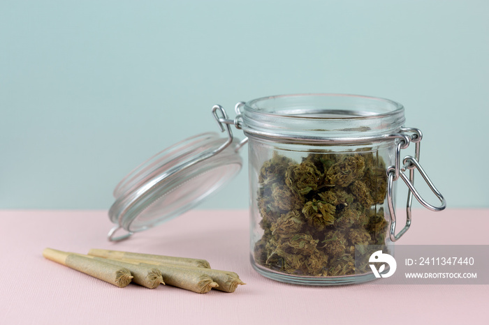 Marijuana in glass jar next to joints arranged on pastel backdrop