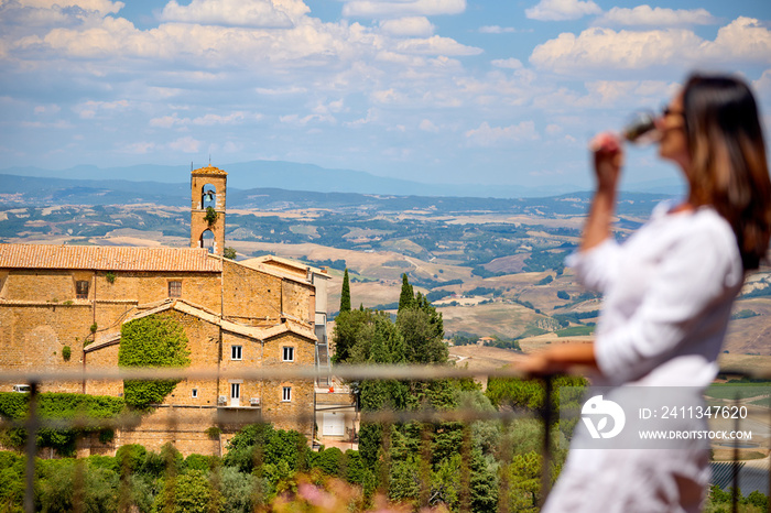 Holidays in Tuscany. Middle aged woman with long hair wearing a white dress watches the landscape of Tuscany from the terrace of a medieval house, enjoying wine. Montalcino, Italy.