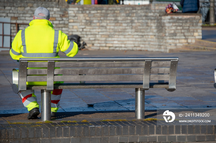 street cleaner wearing hi vis jacket and trousers has a quick coffee or tea break whilst sitting on a bench