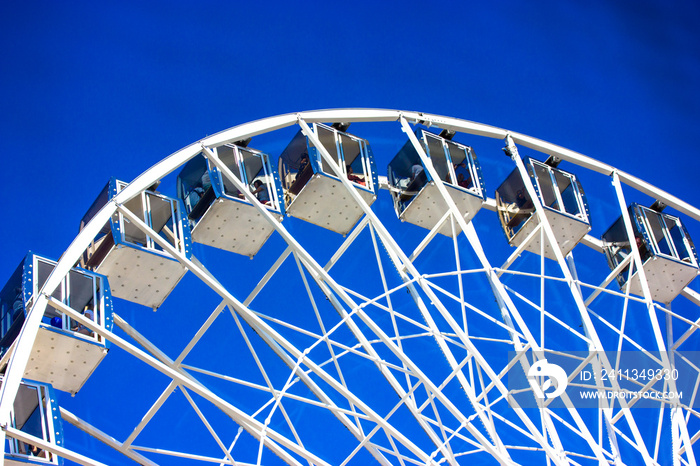 A white Ferris wheel with transparent booths on blue sky background. City entertainment. Luna park games, fun for kids, carrousel. High construction view from below. An attraction for gamblers.