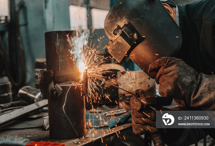 Iron worker in protective suit, mask and gloves welding pipe. Workshop interior.