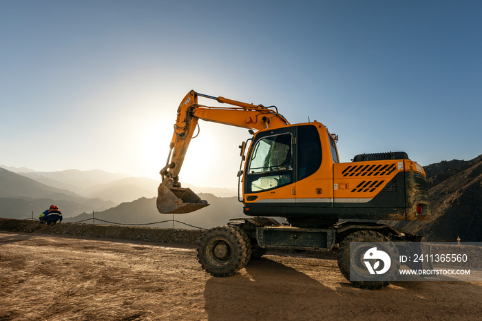 Excavator is working on the hill at sunset. Excavators are heavy construction equipment consisting of a boom, dipper (or stick), bucket and cab on a rotating platform.