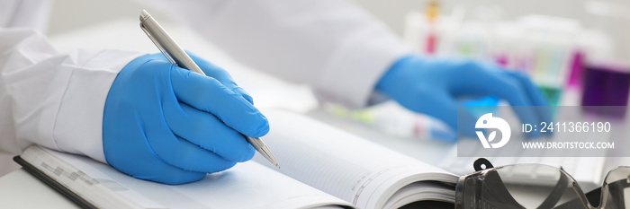 Employee of the chemical laboratory teacher chemist holds a silvery pen in his hand makes notes in the diary records test data from reactions examining test tubes with the substance arm in gloves