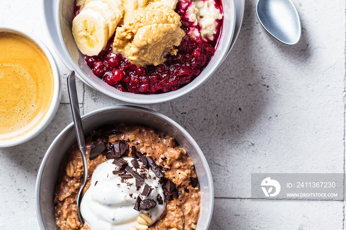 Close up of oatmeal bowls with chocolate, yogurt, fruit and peanut butter, gray tiles background. Healthy breakfast concept.