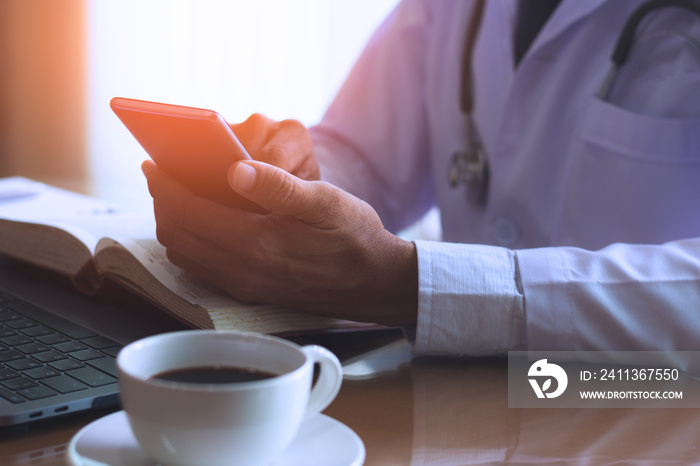 Male doctor hand holding and using mobile smart phone ,work on laptop computer , text book and cup of coffee on the wooden desk in medical room at clinic or hospital.